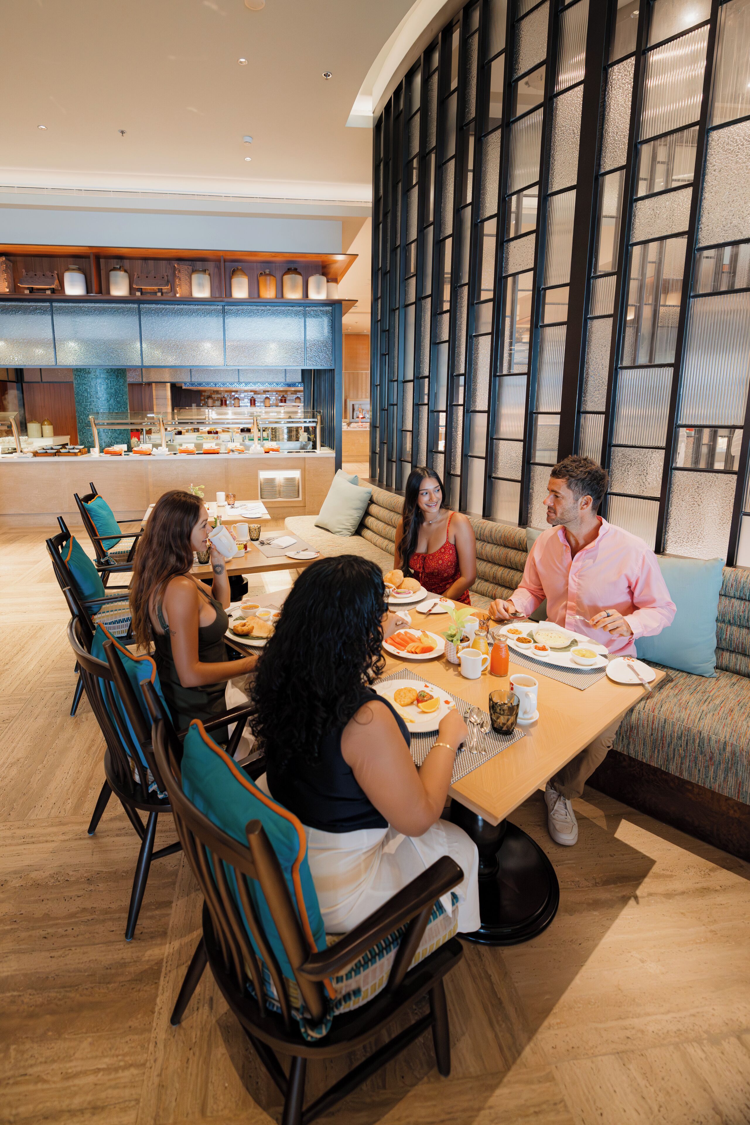 A group of people dining in a modern hotel restaurant with a view of the kitchen and a large window with a grid pattern.