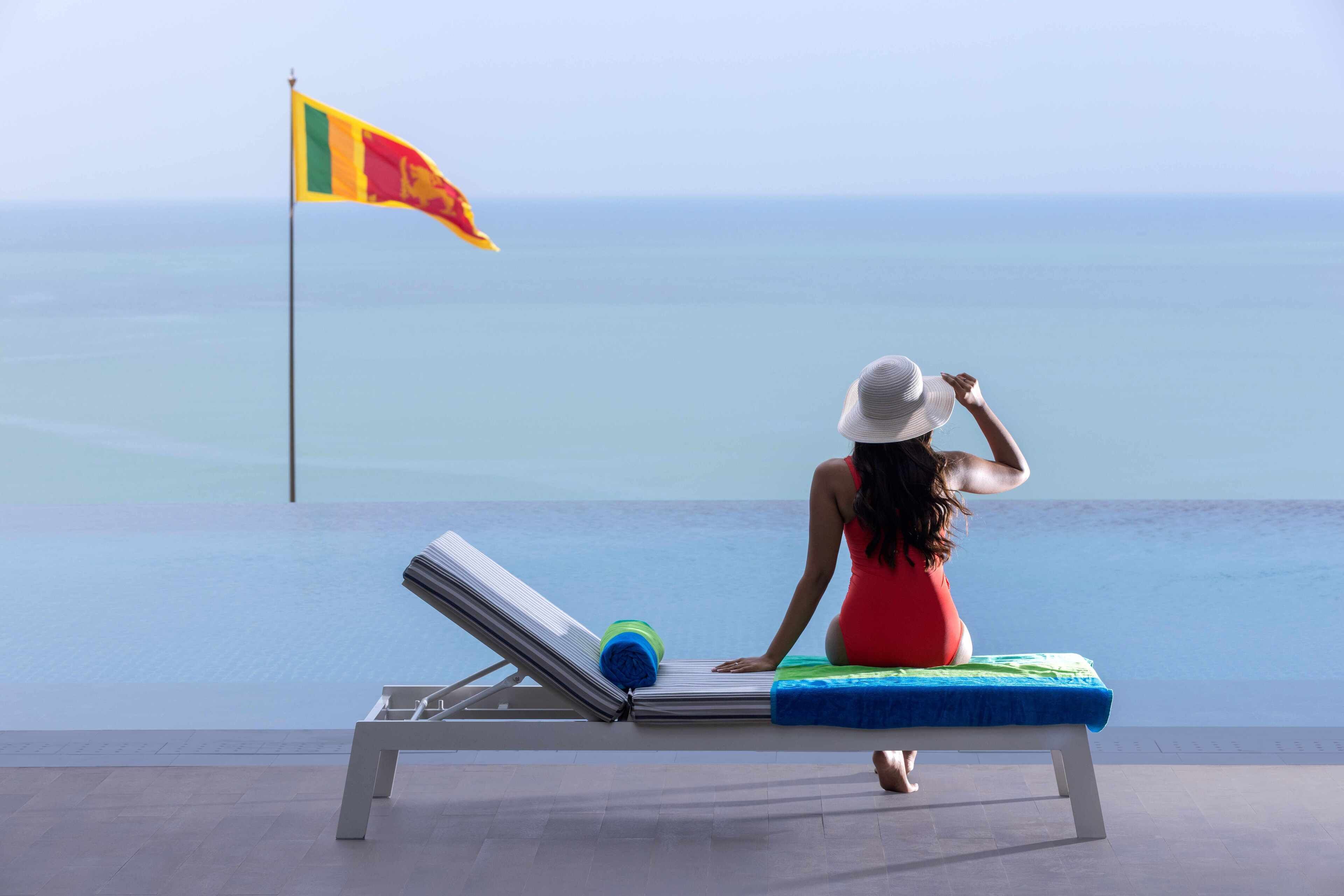 Une femme assise sur une chaise longue, près d’une piscine, avec un drapeau en arrière-plan.