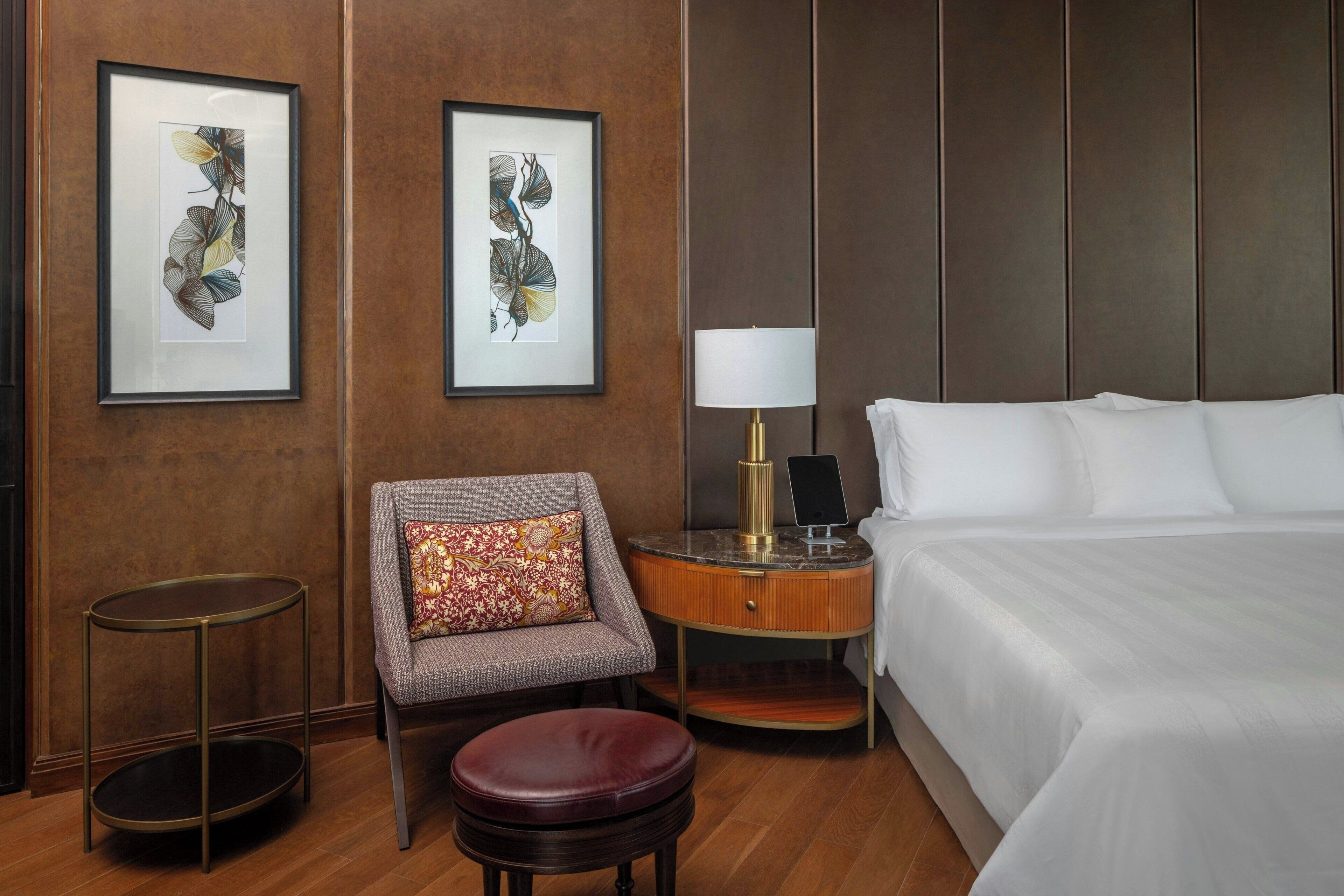 A hotel room with a bed, a chair with a patterned cushion, a small round table with a lamp, and two framed artworks on the wall.