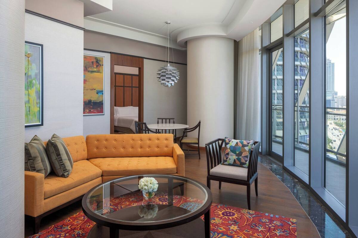 A modern living room with a sofa, a round coffee table with a vase of flowers, a dining area with a table and chairs, and large windows offering a city view.