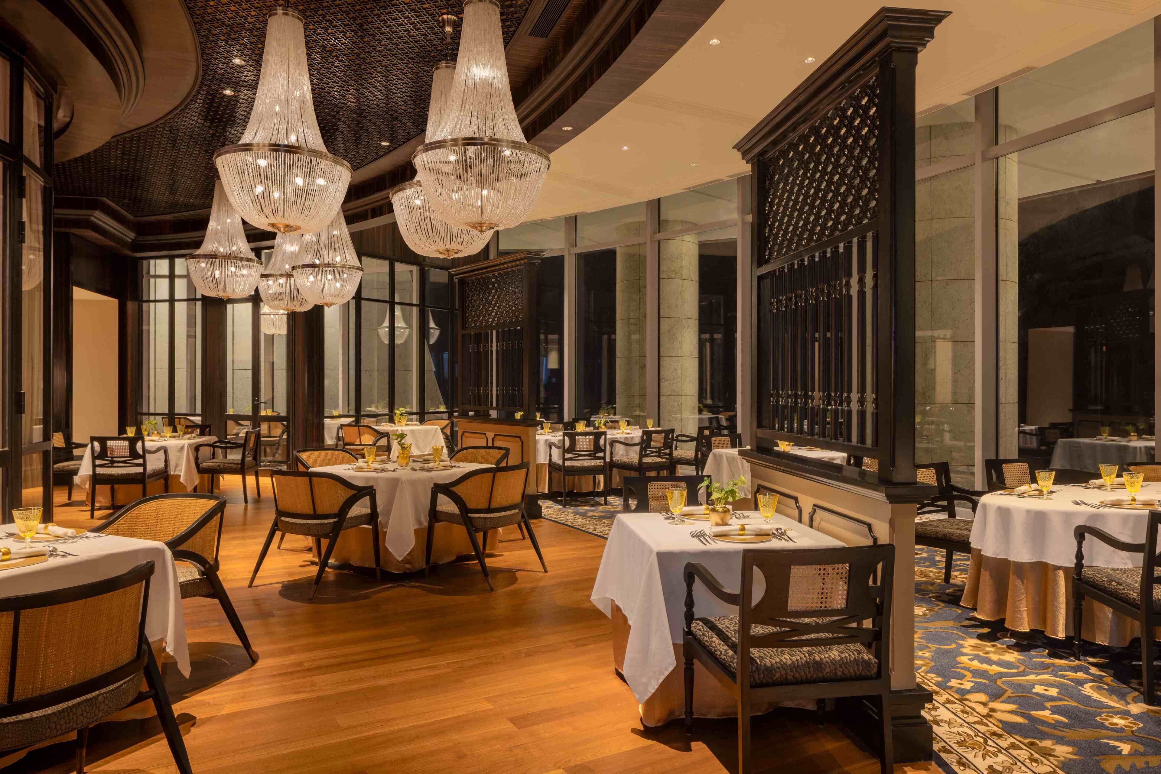 A spacious dining area with multiple tables and chairs, elegant chandeliers, and large windows providing natural light.