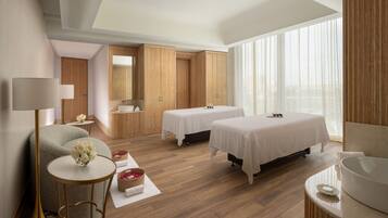 Habitación de hotel moderna con piso de madera, dos camas, un ventanal con cortinas translúcidas, una mesa redonda con un florero y un espejo grande.