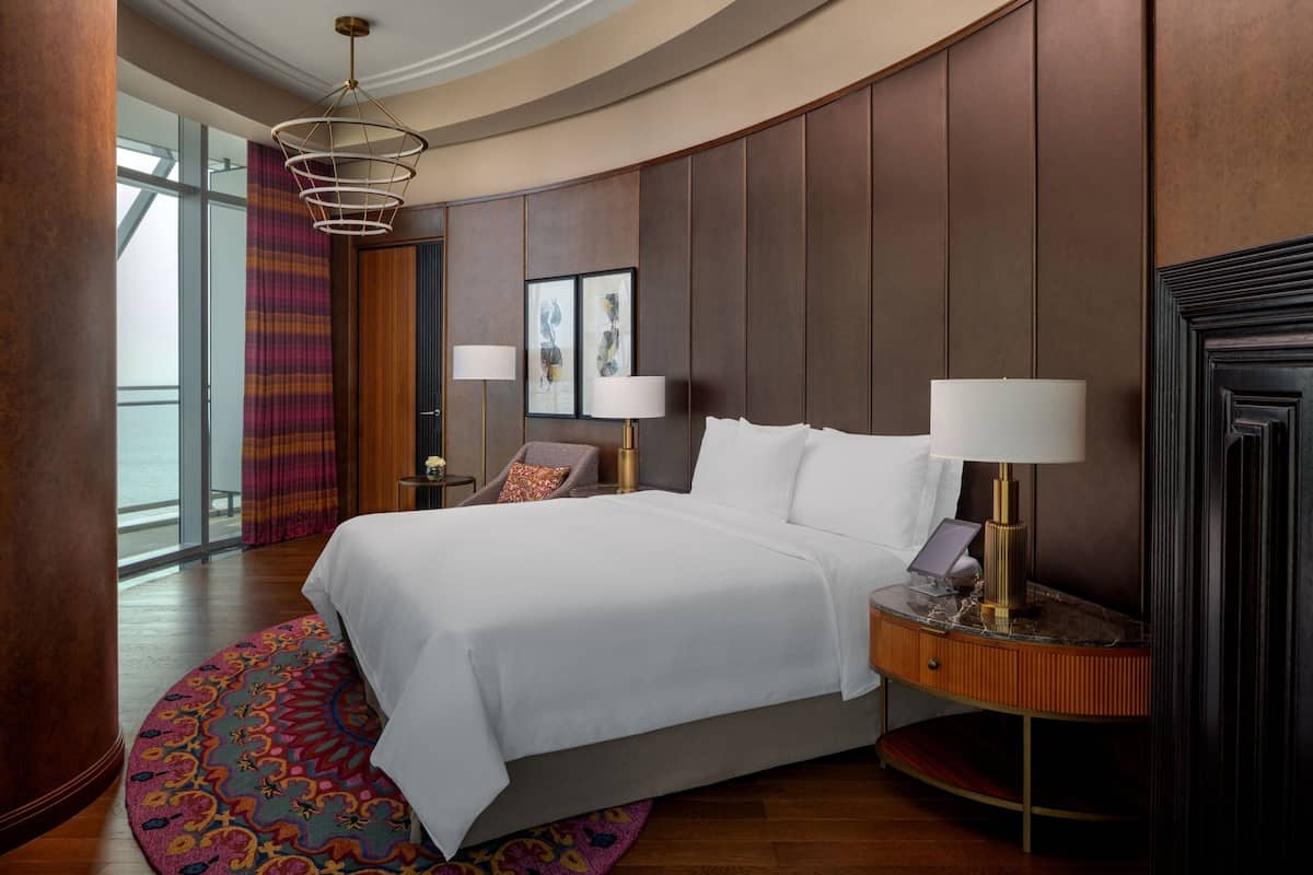 A hotel room with a large bed, two bedside lamps, a wooden headboard, a large window, and a view of the sea.