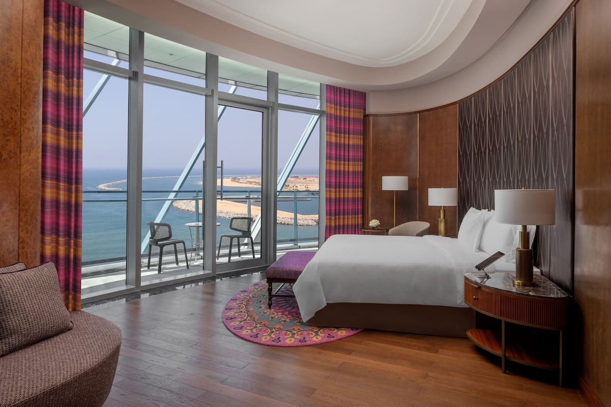 A hotel room with a large window offering a view of a beach and a bridge, a bed, a nightstand with lamps, and a chair.