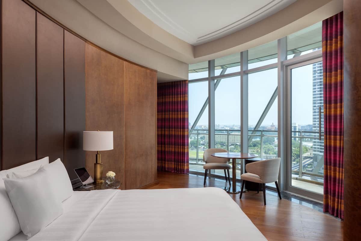 A hotel room with a large bed, a desk with a lamp, a chair, and a view of the city through large windows.