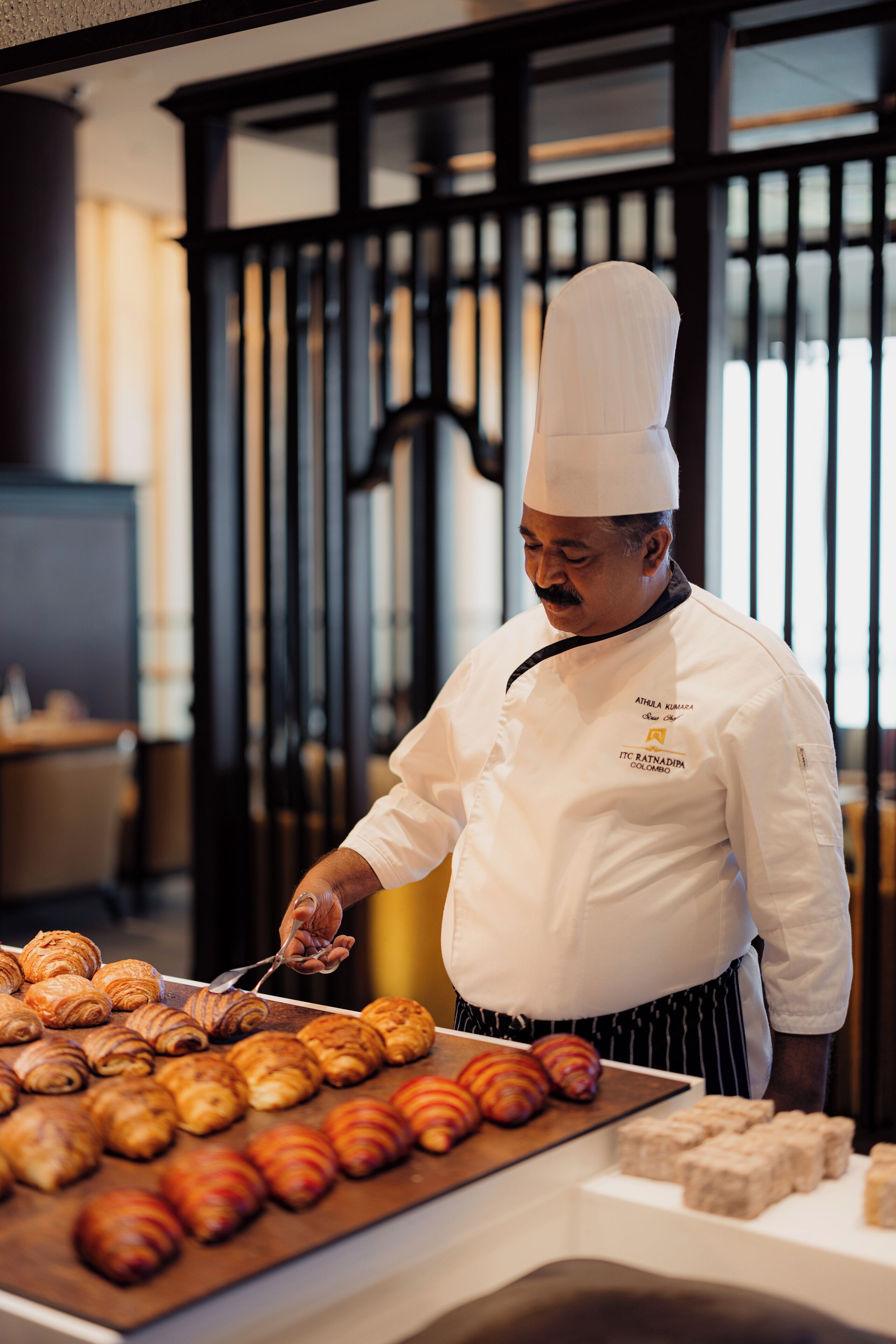A chef in a white uniform with a black apron, standing in a kitchen with a variety of pastries on display.