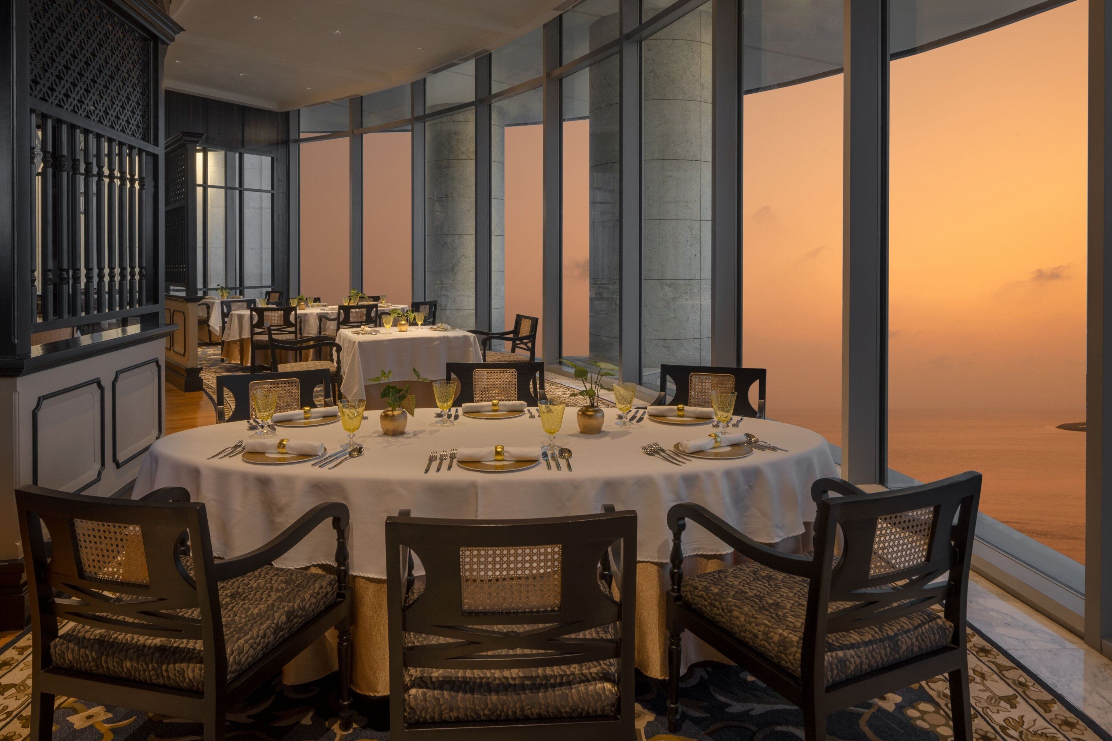 A dining area with round tables set for four, featuring white tablecloths and gold-rimmed plates. The room has large windows offering a view of the sunset.