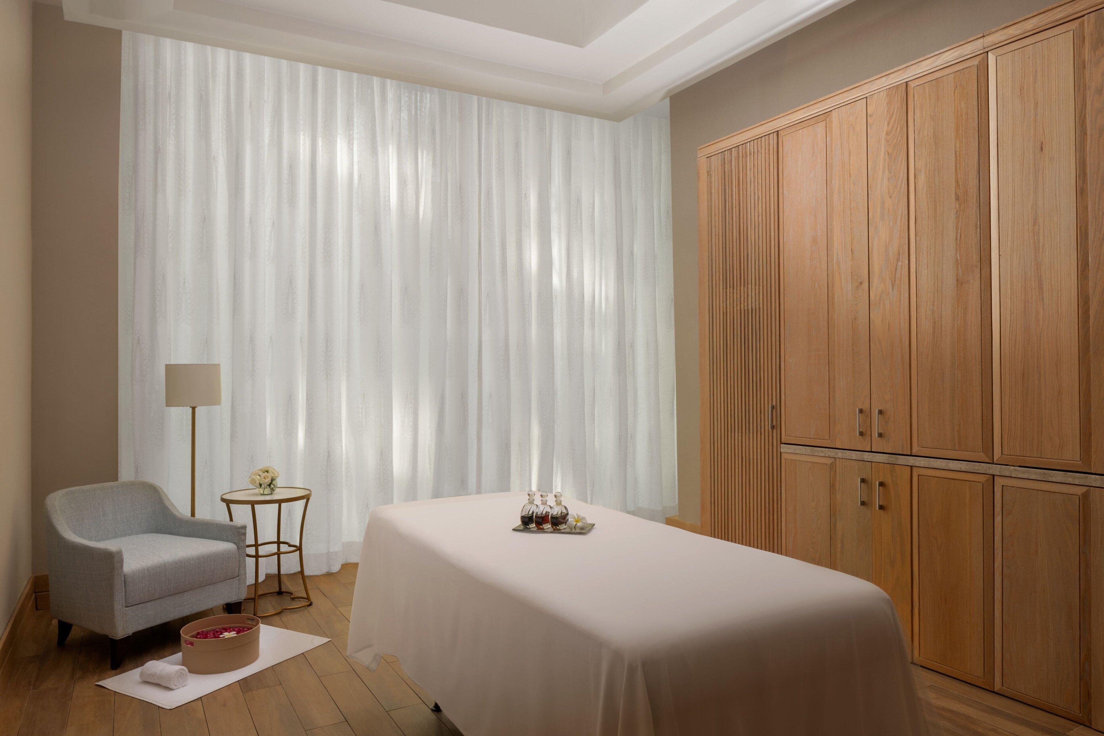 A hotel room with a bed, a chair, a small table with a lamp, and a large wooden wardrobe.