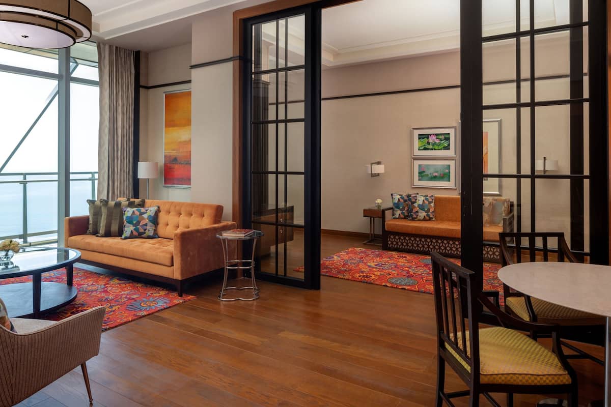 A modern hotel room with a large window, a sofa, a dining table, and a small side table.