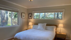 A bedroom with a large bed, two bedside tables, and two lamps. There are two windows providing natural light and a view of trees outside. -  Gracious 3-bedroom Pebble Beach Home in Quiet, Convenient Wooded Area (Del Monte Forest)