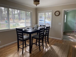 A dining room with a wooden table set for four, flanked by two windows with blinds, a clock on the wall, and a view of the outside. -  Gracious 3-bedroom Pebble Beach Home in Quiet, Convenient Wooded Area (Del Monte Forest)