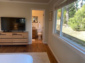 A bedroom with a TV, wooden dresser, and a large window with a view of a backyard. -  Gracious 3-bedroom Pebble Beach Home in Quiet, Convenient Wooded Area (Del Monte Forest)