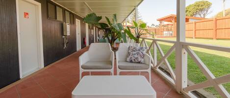 A covered outdoor seating area with two white chairs and a table, a potted plant, and a view of a fenced yard.