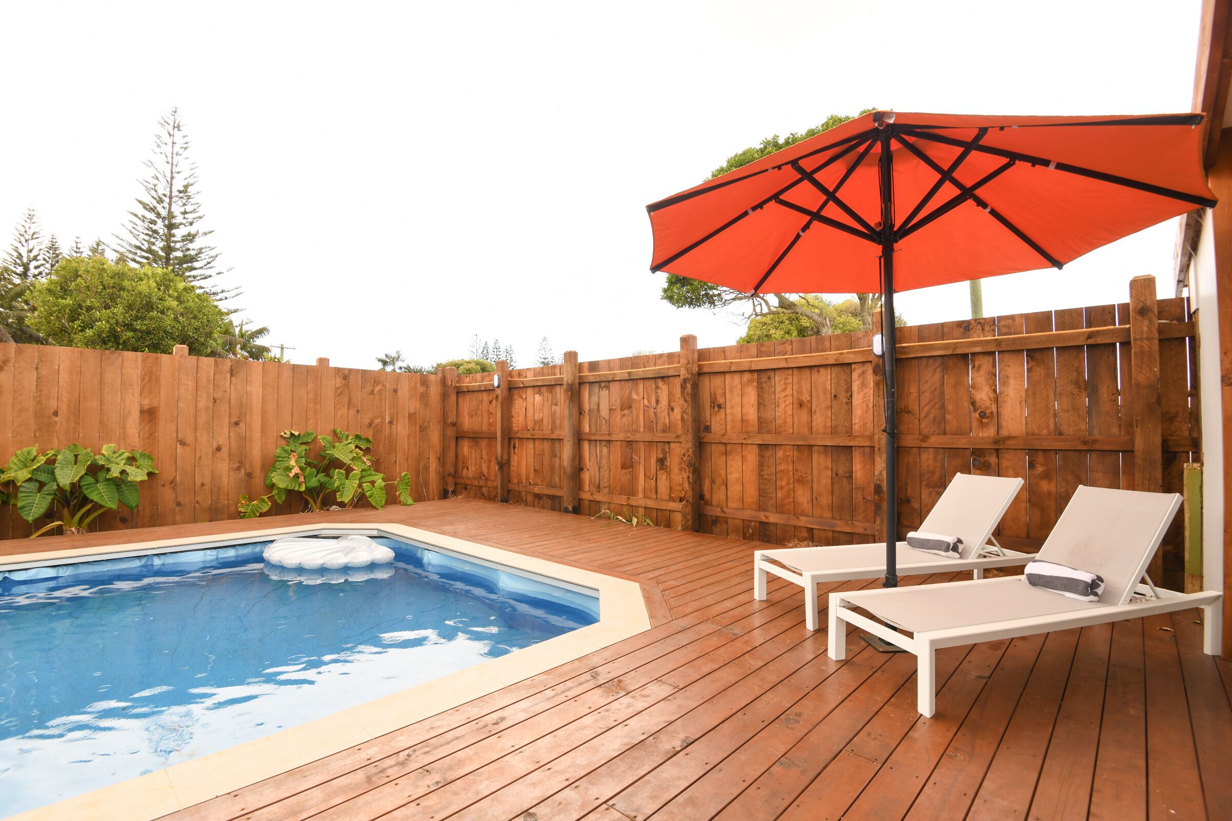 A wooden deck with a swimming pool, two lounge chairs, and a red umbrella.