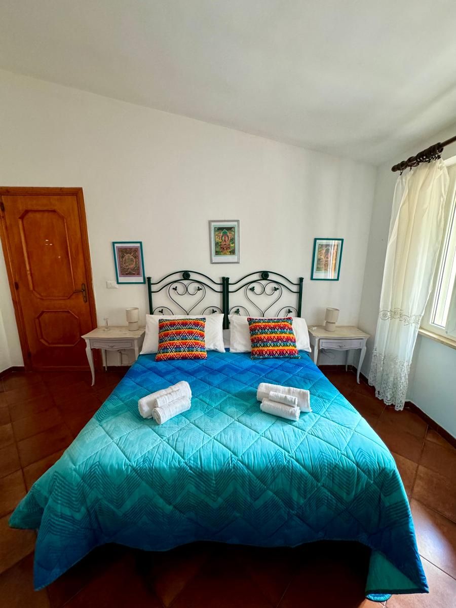 Un lit recouvert d’une couette bleue, deux coussins colorés et des serviettes enroulées posées sur une table de chevet blanche.