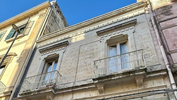 Un edificio histórico con fachada de piedra, un balcón con barandilla de metal y una puerta de madera oscura.