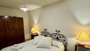 Una cama bien hecha con ropa de cama blanca y toallas enrolladas, un armario de madera, dos mesitas de noche con lámparas y un aplique de techo.