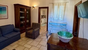 Una sala de estar con un sofá azul oscuro, sillones a cuadros, una mesa de comedor de madera con un tazón verde, y una cocina al fondo.