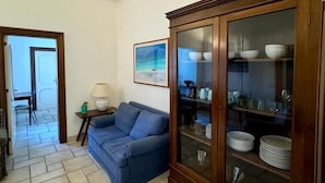 Una sala de estar con un sofá azul, un aparador de madera con puertas de cristal, un cuadro en la pared y suelo de baldosas.