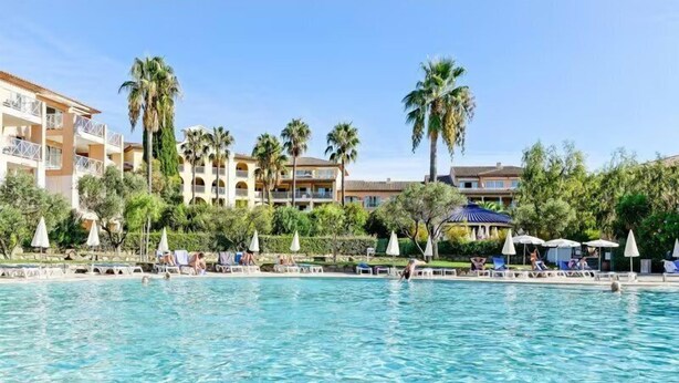 A pool area with lounge chairs and umbrellas, surrounded by palm trees and hotel buildings.