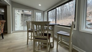 A dining area with a stone exterior wall, large windows, and a view of a wooded area. - Entire Mountain Cottage Near Scenic Hiking Trails, Your Getaway Retreat! (Smithsburg)