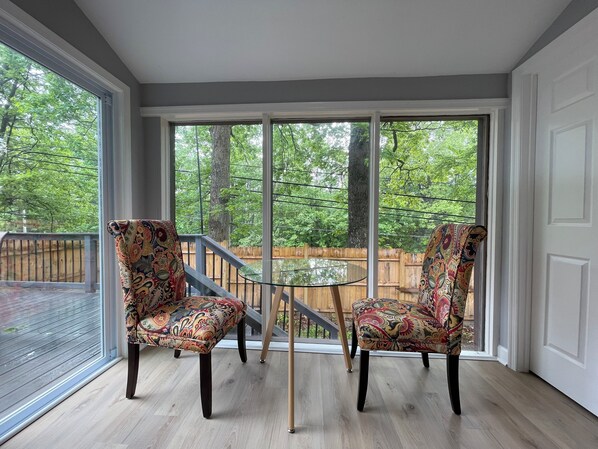 Una habitación con dos sillas estampadas y una pequeña mesa de vidrio, ventanas amplias con vista a árboles y una terraza de madera afuera.