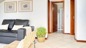 A modern living room with a black leather sofa, a wicker chair, and a potted plant. There are two framed pictures on the wall and a wooden door leading to another room. - Cantinho da Mikaela, a Home in Madeira (Ponta do Sol)