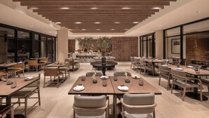 Un vaste espace dédié à la salle à manger, avec de nombreuses tables et chaises, une grande table ronde au centre et une grande fenêtre offrant une vue sur l’extérieur.