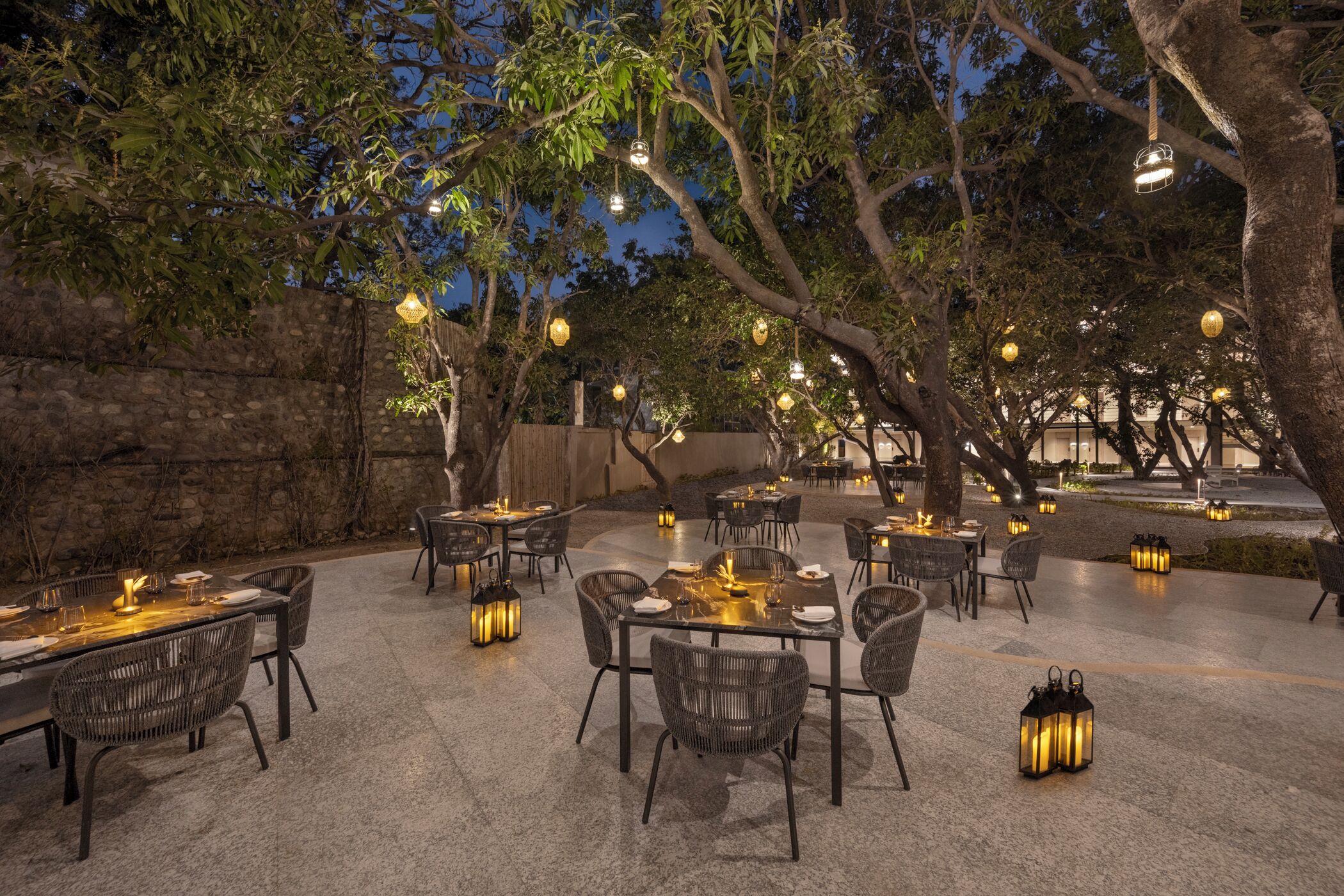 An outdoor dining area with tables and chairs, surrounded by trees and stone walls, with hanging lanterns providing ambient lighting.