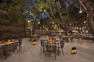 Un área de comedor al aire libre con mesas y sillas, rodeada de árboles y muros de piedra, con faroles colgantes que ofrecen iluminación ambiental.