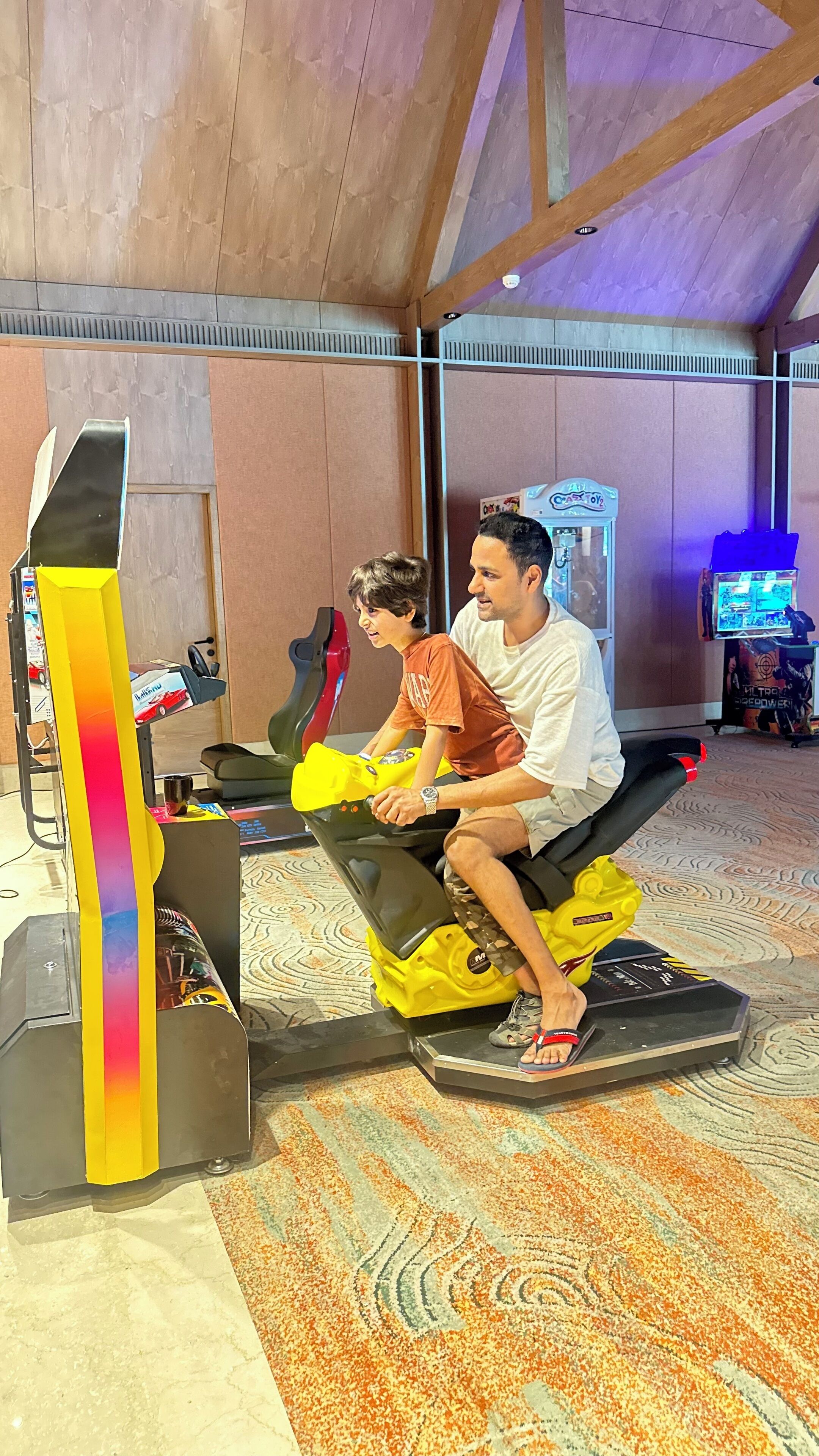 A man and a child on a stationary bike in a room with a patterned carpet and a large poster in the background.