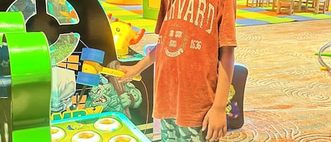 A child standing next to a green arcade machine in a room with wooden beams on the ceiling and colorful play mats on the floor.