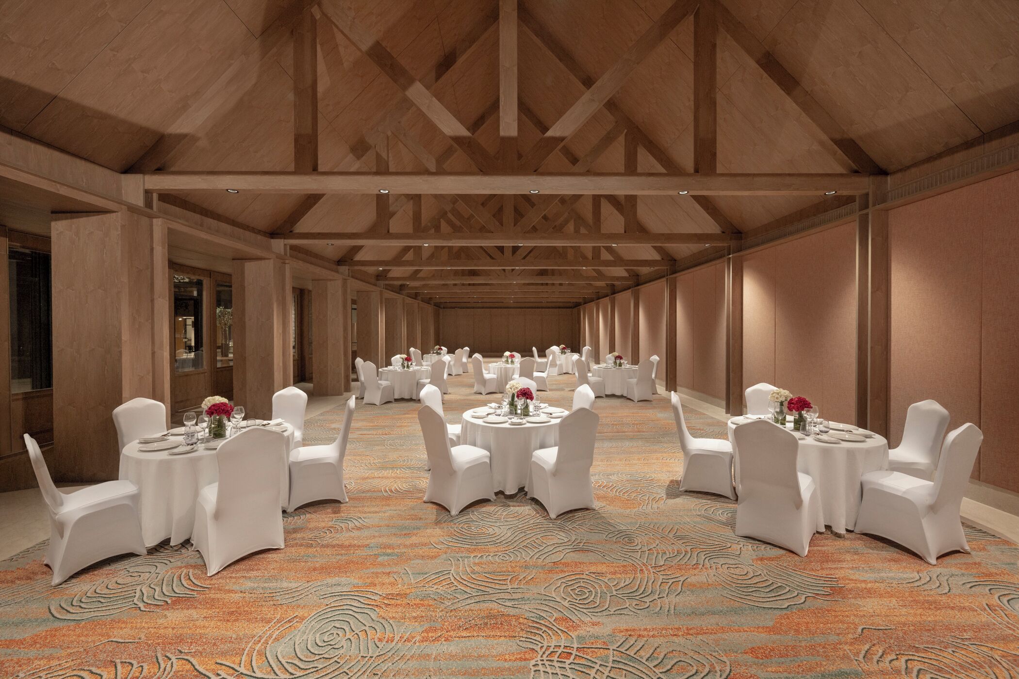 Un salón de banquetes con mesas redondas y sillas blancas, vigas de madera en el techo y una alfombra estampada.