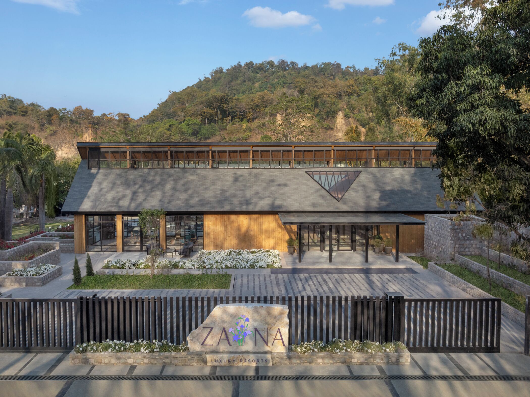 A modern two-story building with a flat roof and large windows, surrounded by landscaped gardens and a mountainous backdrop.