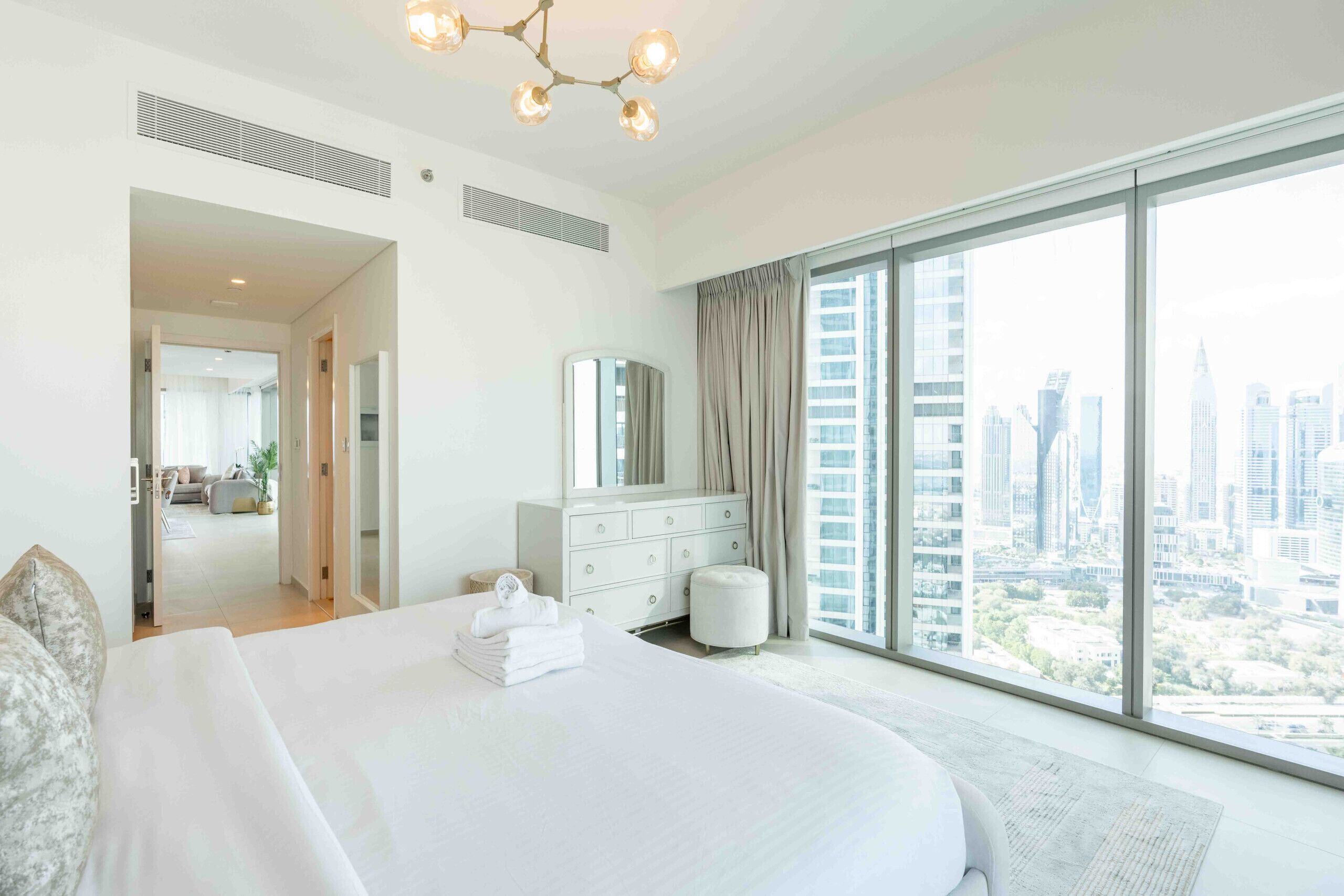 Ein modernes Hotelzimmer mit einem großen Bett, einem Schminktisch und Blick auf eine Skyline durch raumhohe Fenster.