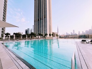 Ein Dachpool mit Liegestühlen und Sonnenschirmen, umgeben von Hochhäusern und wolkenlosem Himmel.