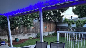 A covered patio with a glass-top table and cushioned chairs, surrounded by a wooden deck and a white railing. - The Tangerine Door (Lewiston)