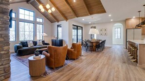 Un vaste espace de vie comprenant un canapé sombre, une table basse ronde et une table à manger avec des chaises. La pièce est dotée d’un plancher en bois, de grandes fenêtres et d’un plafond haut avec des poutres apparentes.
