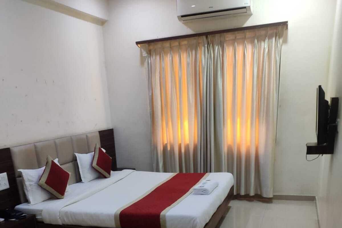 Habitación de hotel con cama, mesita de noche, unidad de aire acondicionado, televisor y ventana con cortinas.