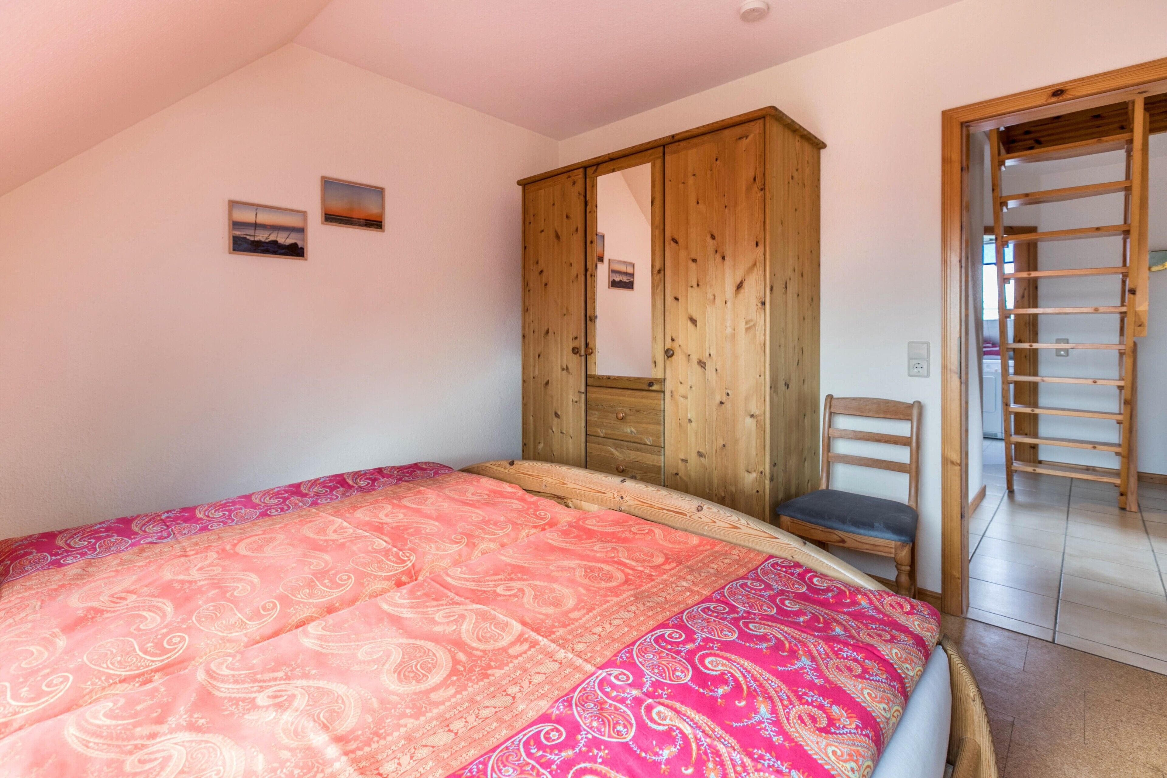 A bedroom with a bed, a wooden wardrobe, a chair, and a ladder leading to a loft.