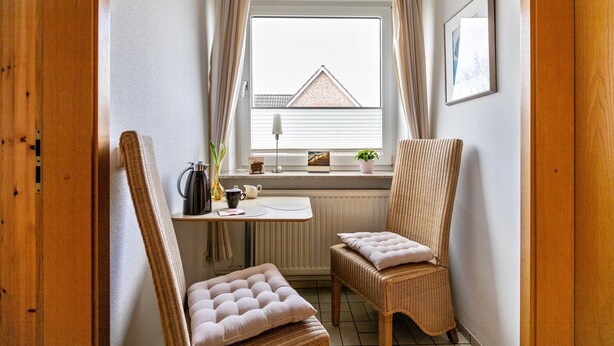 Ein gemütliches Zimmer mit einem großen Fenster, einem Heizkörper, einem Korbstuhl, einem kleinen Tisch und einem gerahmten Bild an der Wand.