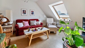 Ein gemütliches Wohnzimmer mit einem roten Sofa, einem Holztisch, einem weißen Sessel und Blick durch eine Dachfenster.