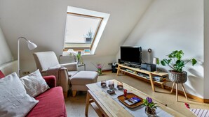 Ein Schlafzimmer im Dachgeschoss mit schrägen Wänden, einem Fernseher, einem Schreibtisch, einem Stuhl, einem Schlafsofa und einem Blumentopf.