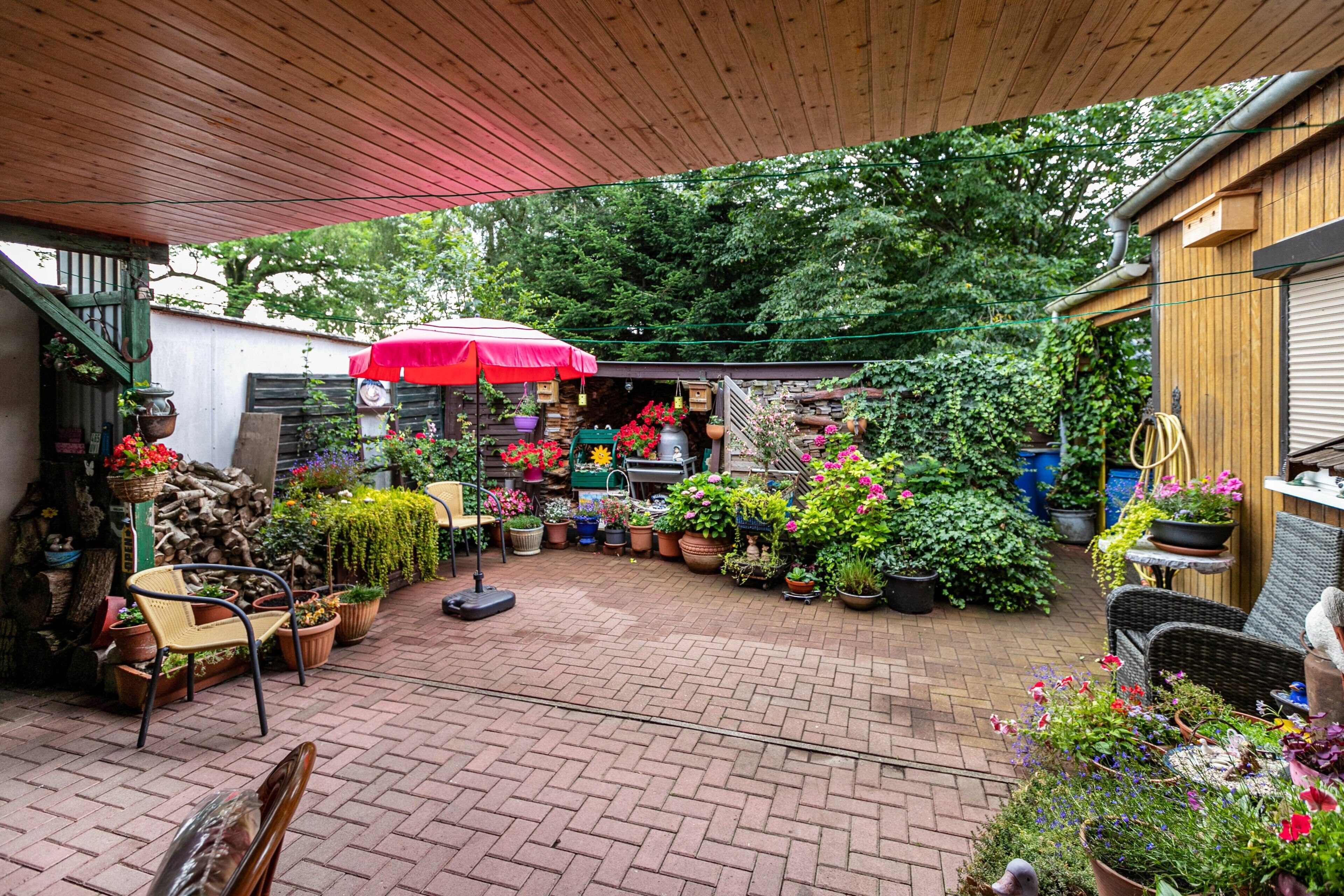 Een terras met plantenpotten, een rode parasol en tuinmeubilair.