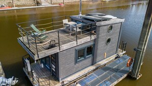 Ein schwimmendes Hausboot mit Deck, Sitzgelegenheiten und einem kleinen Fenster.