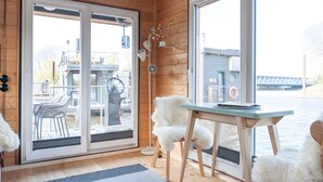 A room with wooden walls, a sliding glass door, a white furry chair, a small table, and a view of a dock and water. - Holiday Home 'Besonderes Tiny Hausboot Marianne' with Private Terrace and Wi-Fi (Hamburg)