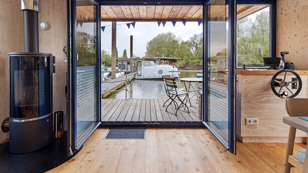 Una cabaña moderna con interior de madera, una puerta de cristal que da a un muelle, un estufa de leña y una mesa con una silla.