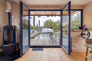 Une cabane moderne avec un intérieur en bois, une porte vitrée donnant sur un quai, un poêle à bois et une table avec une chaise.