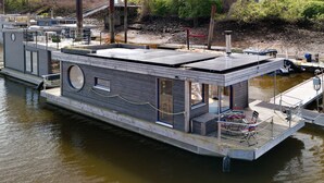 Una casa flotante con terraza, zona de estar techada al aire libre y una pequeña embarcación amarrada en la parte trasera.