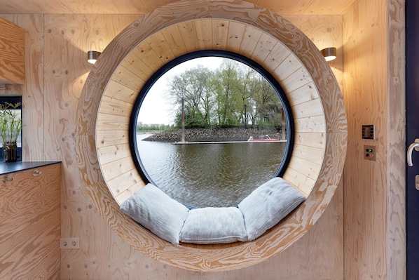 A circular window with a bench inside, wooden walls, and a view of trees and water.